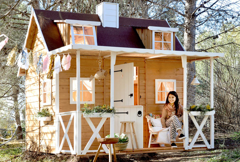 Une jeune fille s'assoit élégamment sur le porche d'une luxueuse maisonnette en bois destinée aux enfants